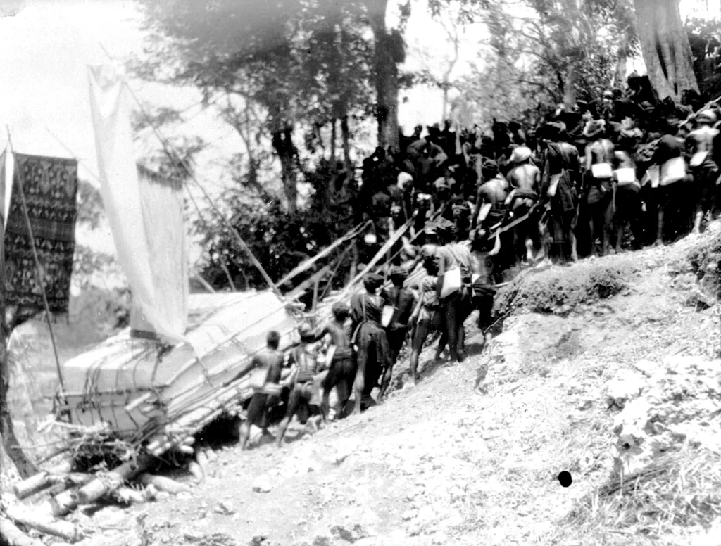 Black and white image shows a large group of people pulling a large structure with sails up a hill in a wooded area. History of Sumba.
