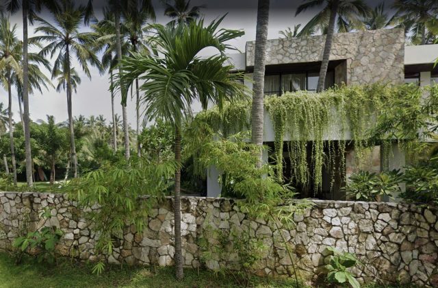 A modern stone and white building with cascading green vines stands amidst tall palm trees and lush tropical foliage, behind a low stone wall.