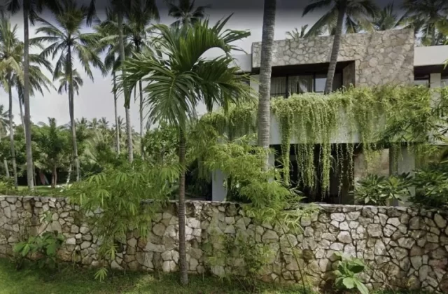 A modern stone and white building with cascading green vines stands amidst tall palm trees and lush tropical foliage, behind a low stone wall.