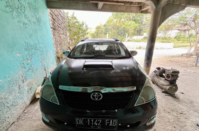 A dark-colored Toyota Kijang Innova is parked under a shelter, with a peeling turquoise wall on the left and green trees in the background.