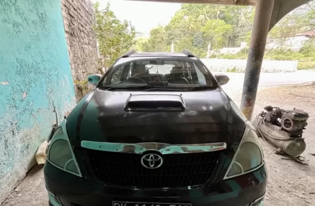 A dark-colored Toyota Kijang Innova is parked under a shelter, with a peeling turquoise wall on the left and green trees in the background.