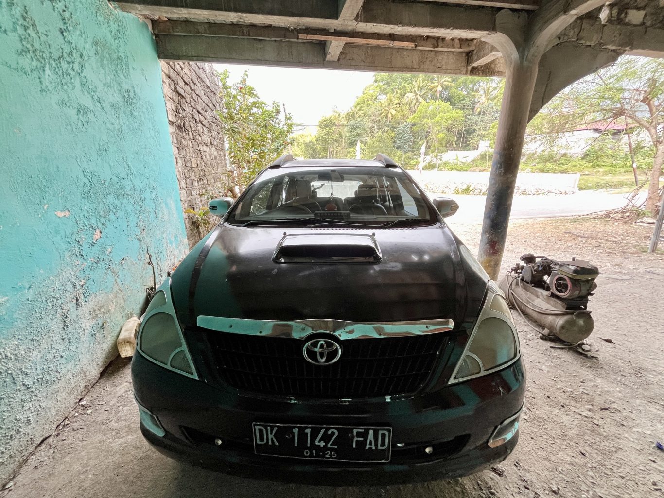 A dark-colored Toyota Kijang Innova is parked under a shelter, with a peeling turquoise wall on the left and green trees in the background.
