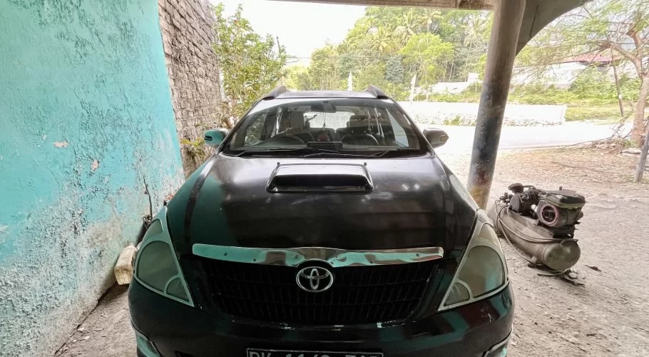 A dark-colored Toyota Kijang Innova is parked under a shelter, with a peeling turquoise wall on the left and green trees in the background.