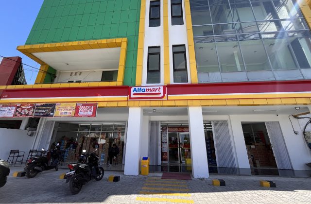 A modern Alfamart mini-market building in Waingapu, Sumba, features green, yellow, white, and red facades with motorcycles parked outside.