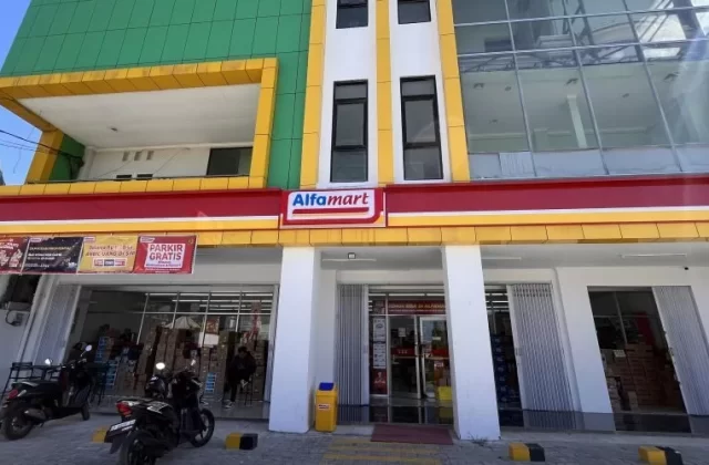 A modern Alfamart mini-market building in Waingapu, Sumba, features green, yellow, white, and red facades with motorcycles parked outside.