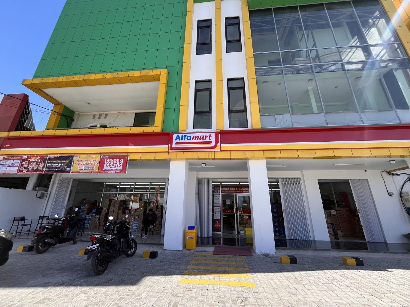 A modern Alfamart mini-market building in Waingapu, Sumba, features green, yellow, white, and red facades with motorcycles parked outside.