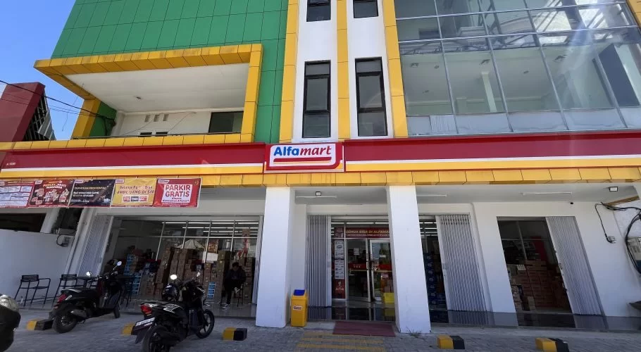 A modern Alfamart mini-market building in Waingapu, Sumba, features green, yellow, white, and red facades with motorcycles parked outside.