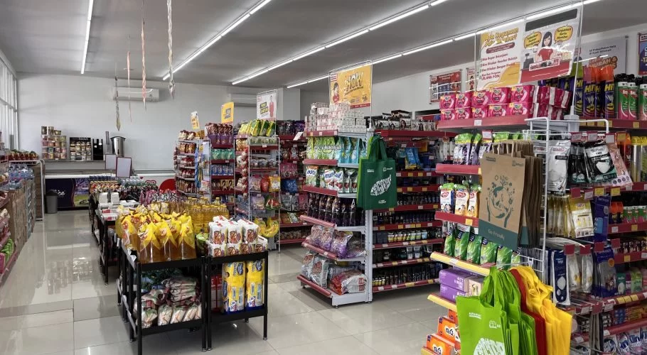 The brightly lit interior of an Alfamart grocery store in Waingapu, Sumba, features multiple aisles filled with colorful packaged food, drinks, and household products on red and white shelves.