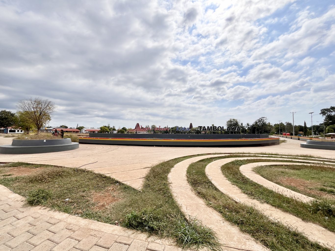 The Alun-Alun Kota Tambolaka city square features a large central sign, paved pathways, and green spaces under a cloudy sky.