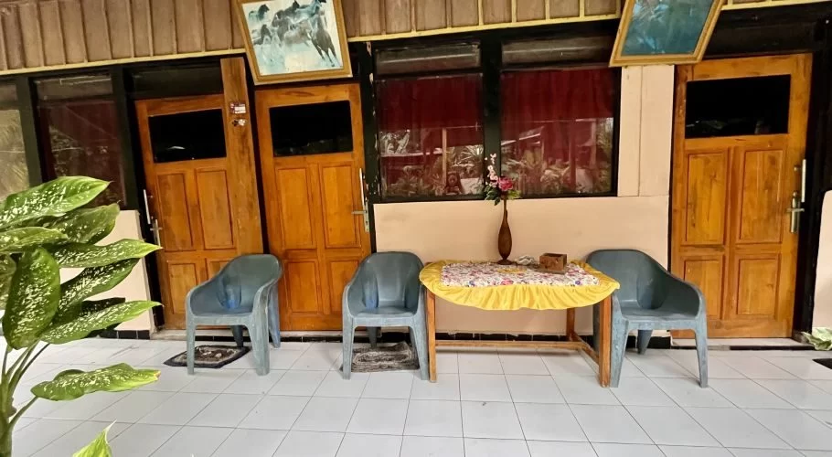 A simple outdoor seating area features a small table with a yellow cloth and three dark grey plastic chairs on a white tiled floor, in front of wooden doors and windows with red curtains.