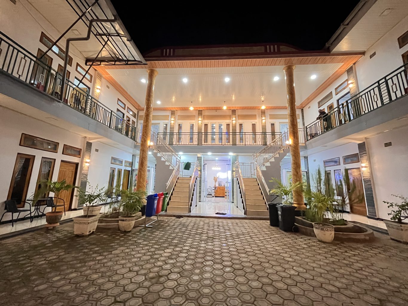 A two-story homestay courtyard at night, brightly lit with warm lights, features symmetrical staircases, balconies, and potted plants.