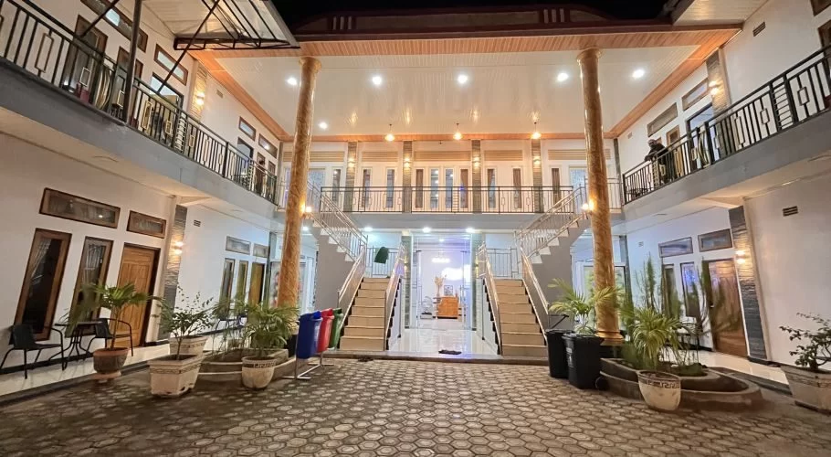 A two-story homestay courtyard at night, brightly lit with warm lights, features symmetrical staircases, balconies, and potted plants.