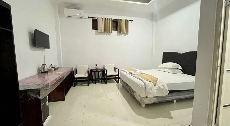A clean, modern hotel room features a bed with white linens, a long desk with a TV, two armchairs, and an air conditioner.