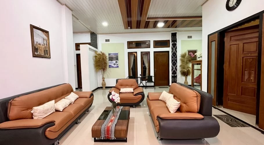 A modern and cozy homestay common area features brown and black sofas, a wooden coffee table, and traditional Indonesian decor.