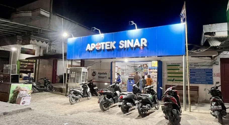 A brightly lit blue pharmacy sign reading "APOTEK SINAR" stands out at night, with several motorbikes parked in front and people visible inside.