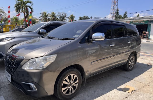 A dark grey Toyota Innova Grand is parked on a sunny street in Waikabubak, Sumba, with a silver SUV behind it and Indonesian flags visible.