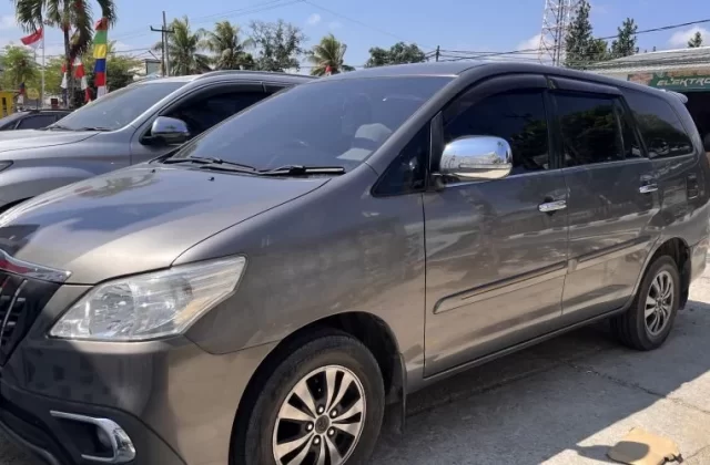 A dark grey Toyota Innova Grand is parked on a sunny street in Waikabubak, Sumba, with a silver SUV behind it and Indonesian flags visible.