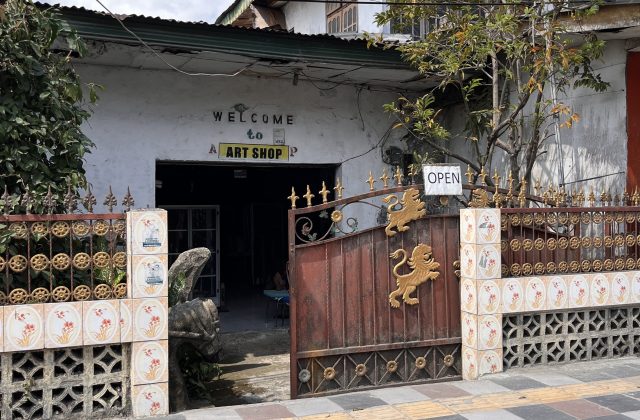 A rustic white building with "WELCOME to ART SHOP" above a dark entrance, partially obscured by a sturdy brown steel gate adorned with golden lions and an "OPEN" sign.