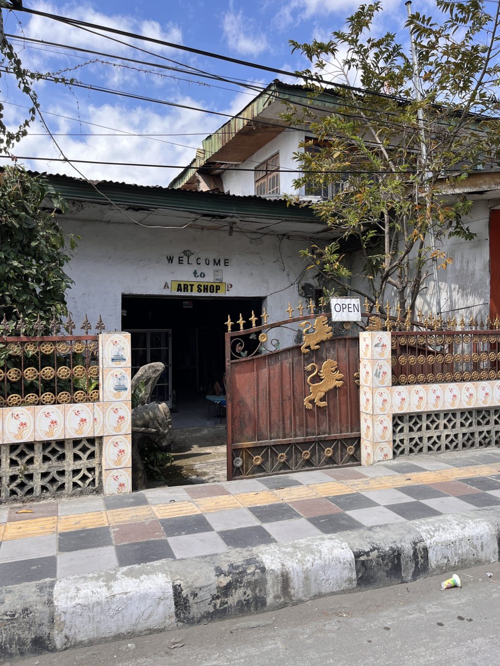 A rustic white building with "WELCOME to ART SHOP" above a dark entrance, partially obscured by a sturdy brown steel gate adorned with golden lions and an "OPEN" sign.
