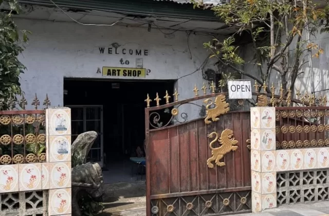 A rustic white building with "WELCOME to ART SHOP" above a dark entrance, partially obscured by a sturdy brown steel gate adorned with golden lions and an "OPEN" sign.