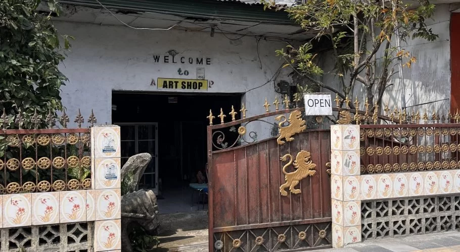 A rustic white building with "WELCOME to ART SHOP" above a dark entrance, partially obscured by a sturdy brown steel gate adorned with golden lions and an "OPEN" sign.
