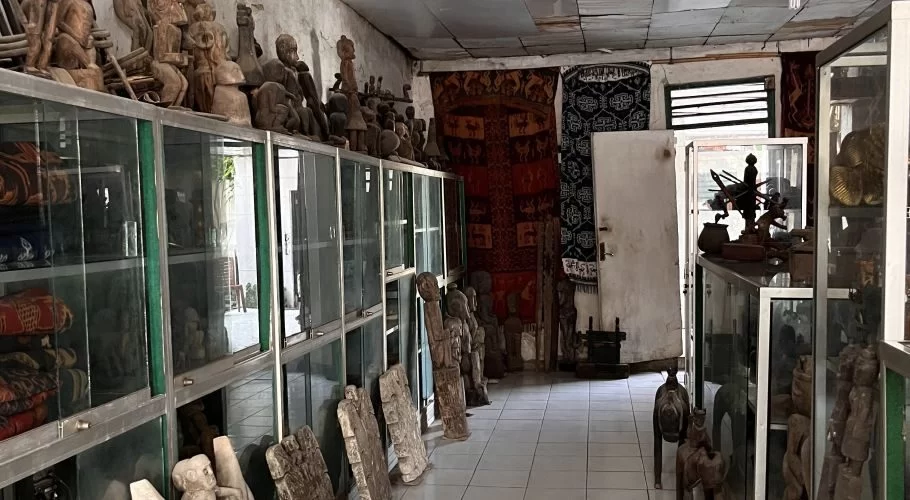 An antique shop interior in Sumba displays numerous wooden carvings on and inside glass cabinets, with colorful patterned textiles hanging on the back wall.