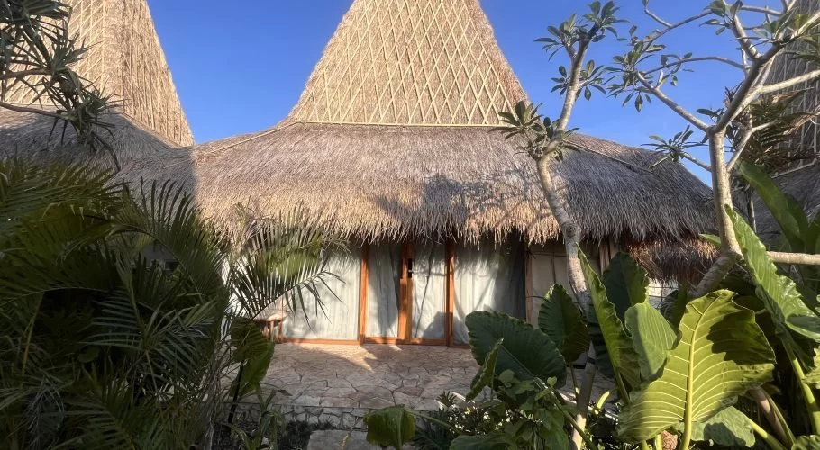 A modern resort villa with a large, conical thatched roof and wooden doors is nestled among lush green tropical plants under a clear blue sky.