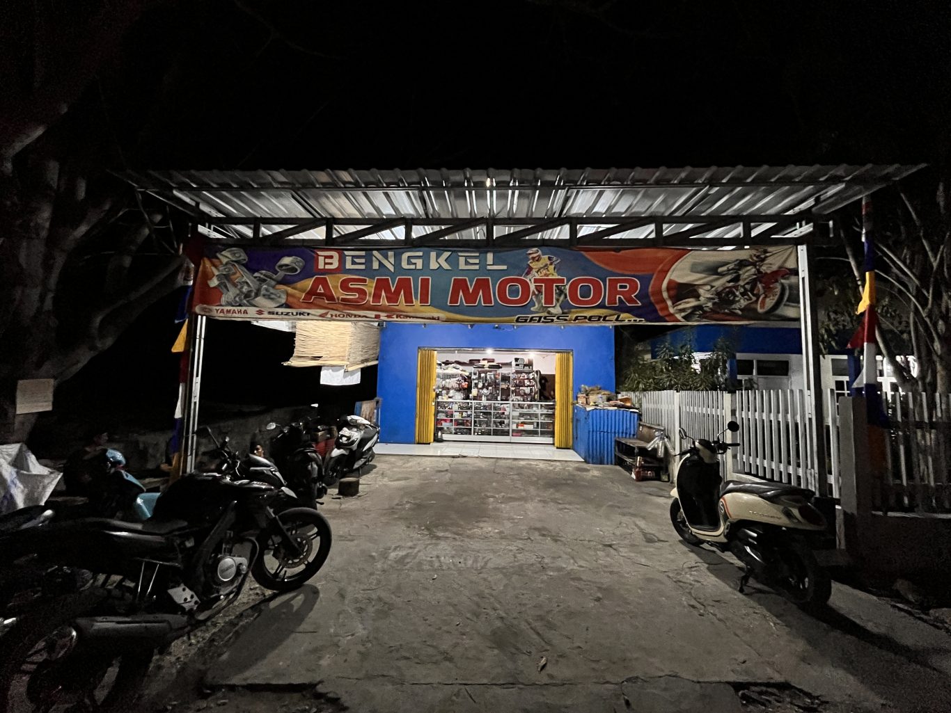 The brightly lit Asmi Motor motorcycle repair shop in Waingapu is open at night, with a blue facade, a large banner, and several motorcycles parked outside.