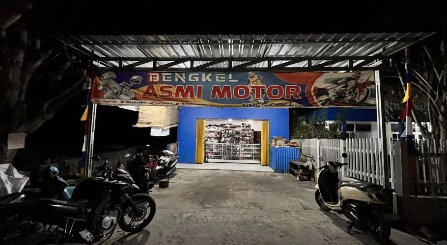 The brightly lit Asmi Motor motorcycle repair shop in Waingapu is open at night, with a blue facade, a large banner, and several motorcycles parked outside.