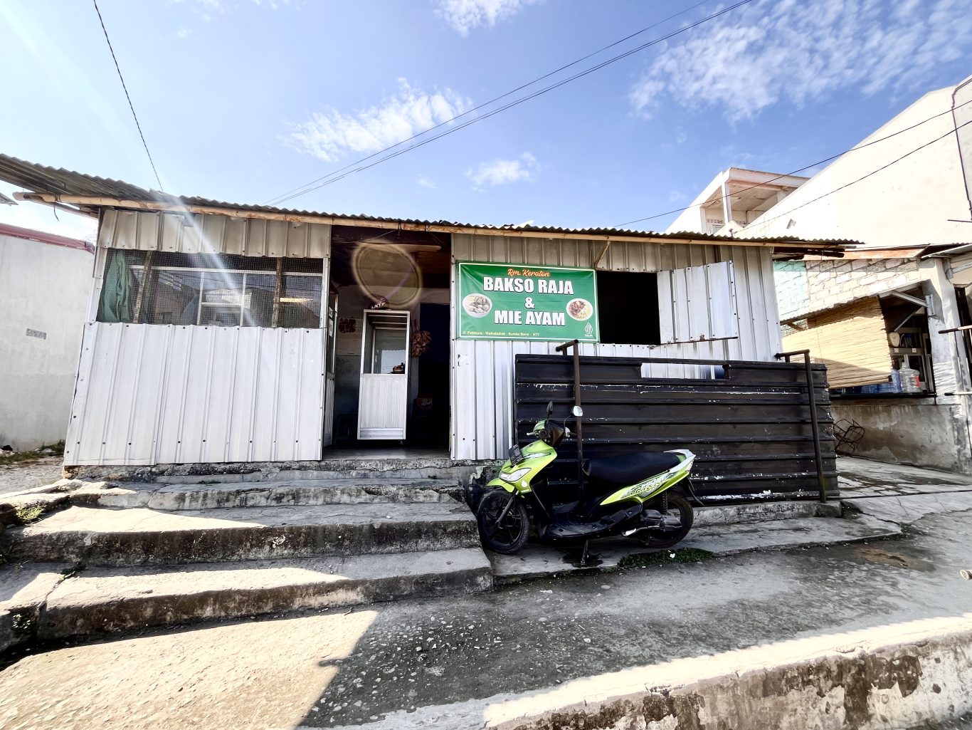 A simple, white corrugated metal building with a green "Bakso Raja & Mie Ayam" sign, and a lime green scooter parked on concrete steps under a blue sky.
