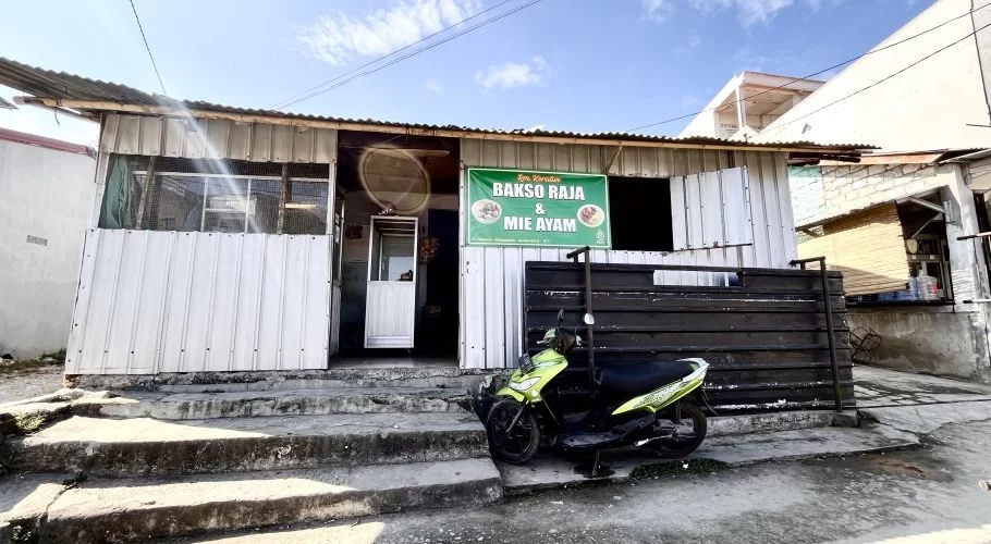 A simple, white corrugated metal building with a green "Bakso Raja & Mie Ayam" sign, and a lime green scooter parked on concrete steps under a blue sky.