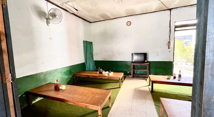 A simple, empty dining room with white walls, a dark green lower section, and light brown wooden tables set with condiments, a wall fan, and a small TV.