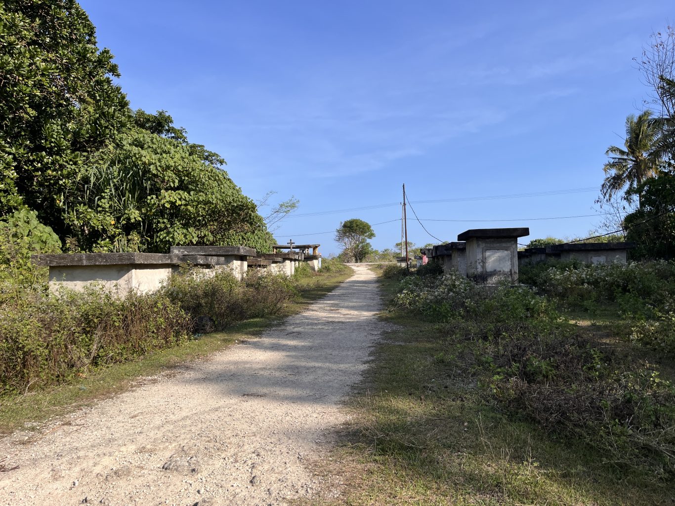 A dirt path leads into Balani Village, flanked by concrete graves and dense green foliage under a clear blue sky.