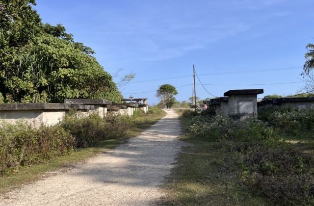 A dirt path leads into Balani Village, flanked by concrete graves and dense green foliage under a clear blue sky.