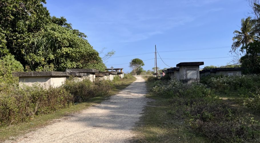 A dirt path leads into Balani Village, flanked by concrete graves and dense green foliage under a clear blue sky.