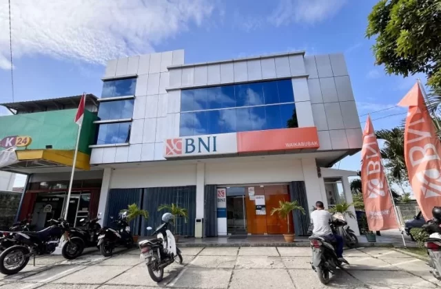 The modern two-story Bank BNI building in Waikabubak features a grey facade, reflective blue windows, and orange branding, with motorbikes parked outside under a cloudy sky.