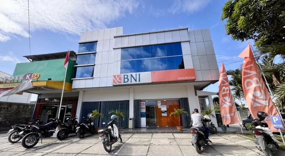 The modern two-story Bank BNI building in Waikabubak features a grey facade, reflective blue windows, and orange branding, with motorbikes parked outside under a cloudy sky.