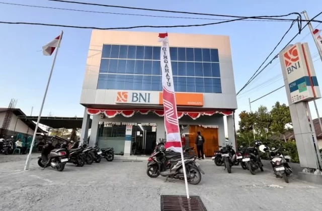 A modern, two-story Bank BNI building with reflective blue windows and orange logos stands under a clear sky, flanked by parked motorcycles and Indonesian flags.
