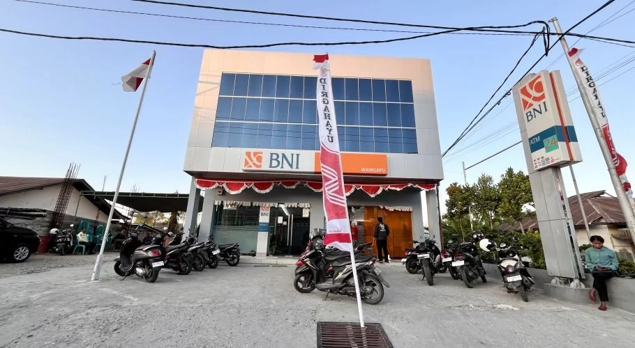 A modern, two-story Bank BNI building with reflective blue windows and orange logos stands under a clear sky, flanked by parked motorcycles and Indonesian flags.