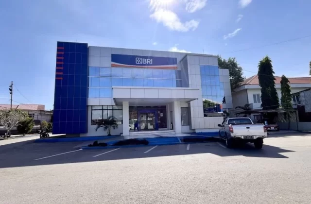 A modern two-story Bank BRI building with blue and silver facade stands under a bright blue sky, with a silver pickup truck parked in front.