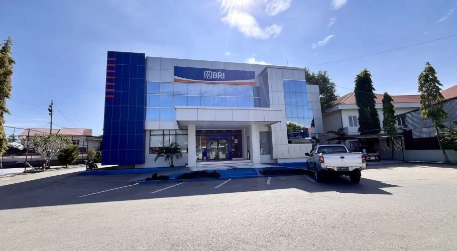 A modern two-story Bank BRI building with blue and silver facade stands under a bright blue sky, with a silver pickup truck parked in front.