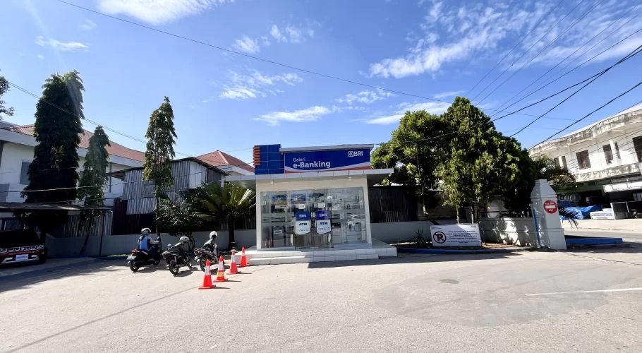 A modern glass-fronted Bank BRI ATM building with four machines stands on a sunny day in Waikabubak, Sumba, with motorcycles parked nearby.