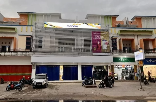 The Bank Mandiri branch in Tambolaka, a multi-story building with colorful accents, stands on a street with parked vehicles and people.