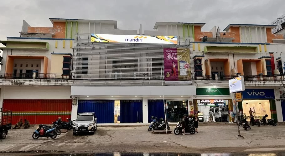 The Bank Mandiri branch in Tambolaka, a multi-story building with colorful accents, stands on a street with parked vehicles and people.