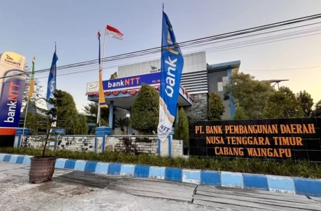 Visible is the Bank NTT Waingapu building, a modern structure with blue and white branding, flags, and a large black sign in front, under a clear sky.
