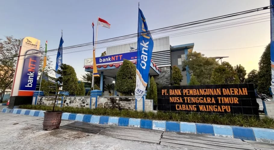 Visible is the Bank NTT Waingapu building, a modern structure with blue and white branding, flags, and a large black sign in front, under a clear sky.