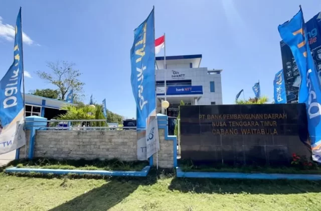 A modern white and blue Bank NTT Waitabula building with a large dark sign and several blue branded flags stands under a clear blue sky.