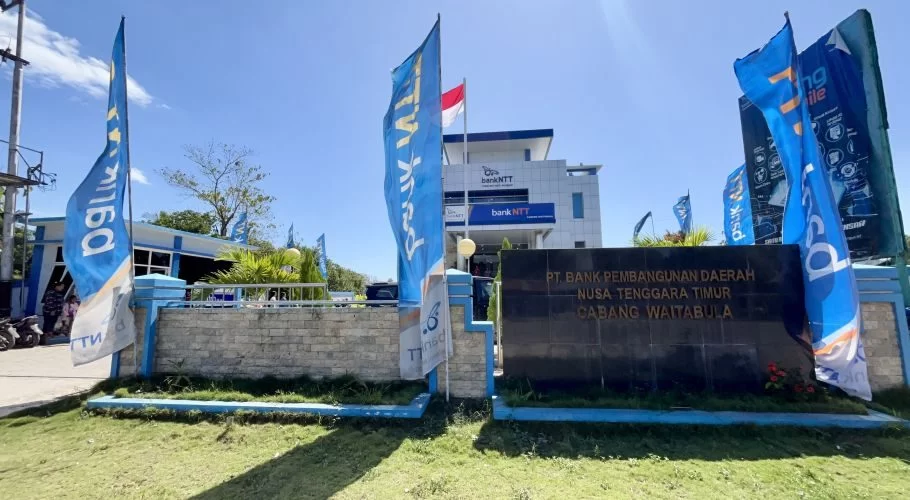 A modern white and blue Bank NTT Waitabula building with a large dark sign and several blue branded flags stands under a clear blue sky.