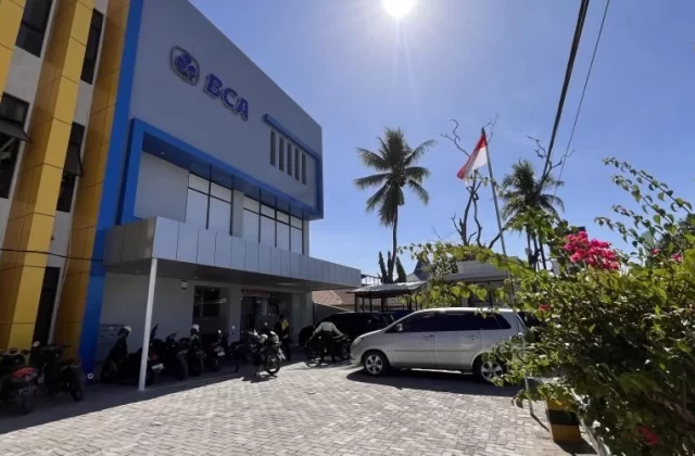 A modern BCA Bank building with blue and yellow accents stands under a bright blue sky, with motorcycles and a silver car parked on a paved lot, alongside palm trees and an Indonesian flag.
