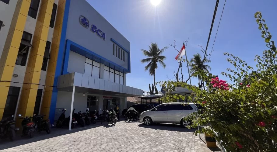 A modern BCA Bank building with blue and yellow accents stands under a bright blue sky, with motorcycles and a silver car parked on a paved lot, alongside palm trees and an Indonesian flag.